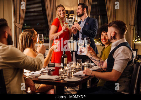 Friends are at a formal meal together. A couple are standing together at one end of the table, making a toast to which everyone is raising their glass Stock Photo