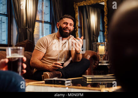 Man is sitting in the lounge of a bar with some friends. He is talking to them with a pint of beer in his hand. Stock Photo