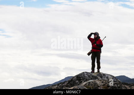 Female tour leader with a rifle stood on high point on look out for Polar Bears to protect tourists visiting Hvalsoy Qaqortoq Kujalleq South Greenland Stock Photo