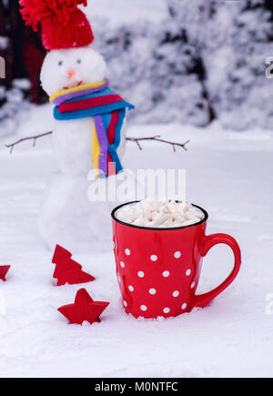 red ceramic mug with white polka dots with hot chocolate and marshmallows, behind a cheerful snowman on snow Stock Photo