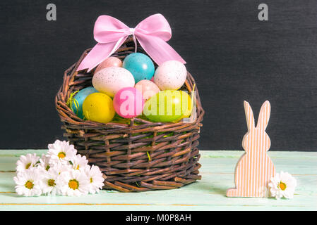 Wicker basket full of multicolored Easter eggs and a pink bow, surrounded by white daisies and a wooden bunny, on a table and a black wall. Stock Photo