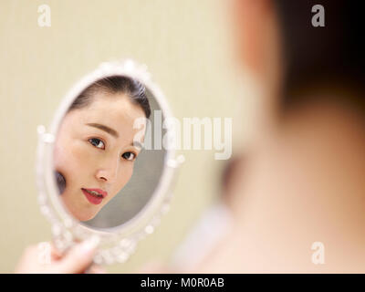 beautiful young asian woman looking at self in mirror while applying make-up on face using brush.