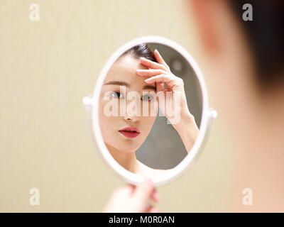 beautiful young asian woman looking at self in mirror, hand on forehead.