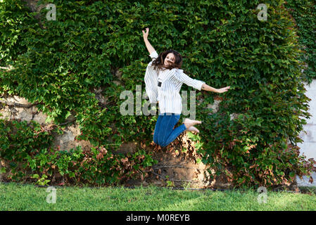 Happy millennial girl jumping freely against garden wall in summer. Stock Photo