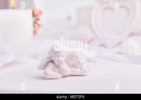 statuette of two antique little lovely angels of the gypsum on the white wooden table with photo frame, candle and flowers. Love background. Copy space, selective focus. Stock Photo