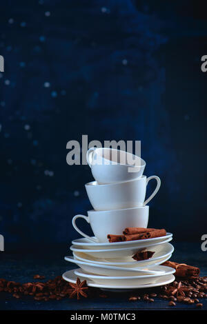 Stack of balancing white coffee cups and tea saucers with coffee beans and cinnamon on a dark blue background. Creative barista concept with copy spac Stock Photo