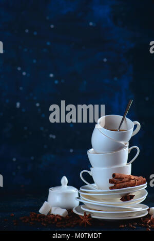 Stack of balancing white coffee cups and tea saucers with coffee beans and cinnamon on a dark blue background. Creative barista concept with copy spac Stock Photo