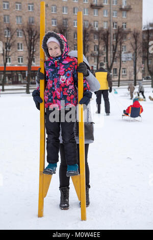 Mom supports from the back of the child standing on wooden stilts at winter. Social activities are at Maslenitsa holiday, Russia Stock Photo