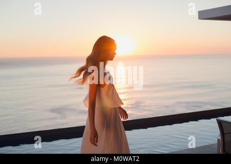 Woman in white dress on tranquil luxury patio with sunset ocean view Stock Photo