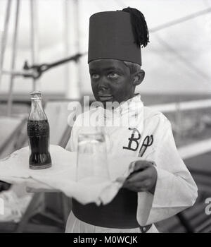 1950s, historical, childrens' fancy dress party onboard a Union-castle steamship going to South Africa....picture shows a young boy carrying a tray with a coca-cola bottle on it and with boot polish on his face and wearing the party uniform and fez (headdress) based on a foreign, possibly, Egyptian, waiter from Cairo. The great British comedian, Tommy Cooper used a fez as part of his comedy/magic act and was said to have had the idea after seeing an Egyptian waiter while serving with the British Army in Cairo. Stock Photo