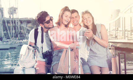 group travellers taking selfie on camera in smartphone on waterfront in town on summer day Stock Photo
