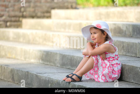 Adorable little girl wearing white hat  sitting on stairs on warm and sunny summer day Stock Photo