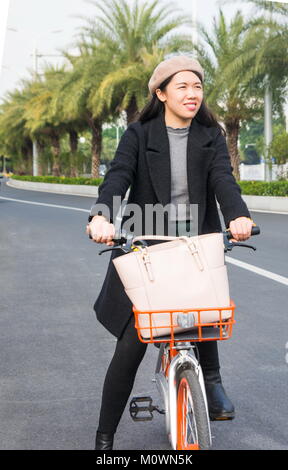 Asian office lady riding public bicycle through urban area Stock Photo