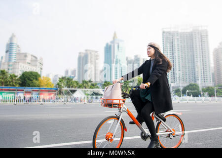 Asian office lady riding public bicycle through urban area Stock Photo