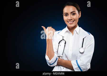 Young happy lady with stethoscope looking camera and pointing at copy space isolated over black Stock Photo