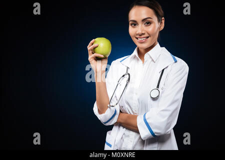 Young smiling lady doctor in medical gown holding green apple and looking camera isolated over black Stock Photo
