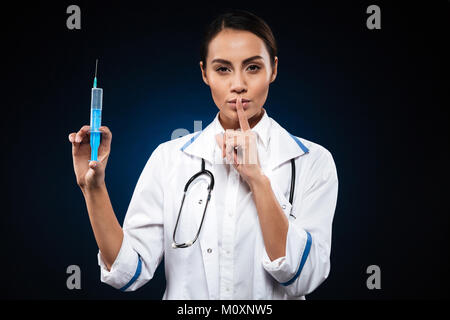 Calm serious lady nurse holding syringe and making silence gesture isolated over black Stock Photo