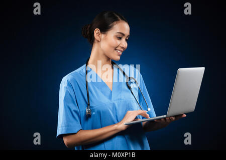 Cheerful lady nurse in medical blue uniform using laptop and looking camera isolated over black Stock Photo