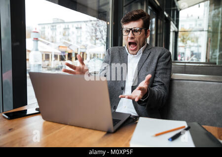 Angry shocked business man in eyeglasses sitting by the table in cafe while looking at laptop computer Stock Photo