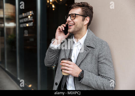 Happy business man in eyeglasses and coat talking by smartphone while holding cup of coffee and looking away outdoors Stock Photo
