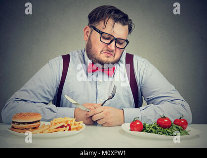 Overweight man in formal outfit looking at salad†with hate while craving juice hamburger. Stock Photo