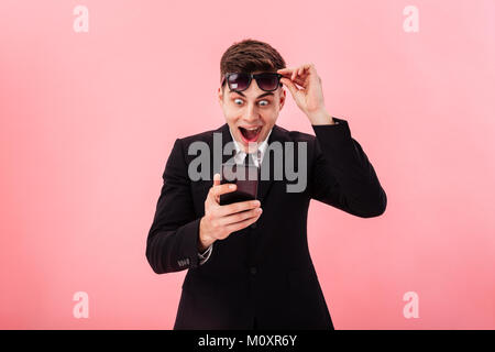 Surprised happy man taking off sunglasses using smartphone isolated over pink Stock Photo
