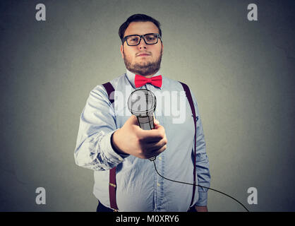 Young chunky man in eyeglasses outstretching microphone at camera looking serious. Stock Photo
