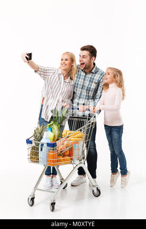 Full length portrait of a happy family standing with a shopping trolley full of groceries and taking a selfie isolated over white background Stock Photo