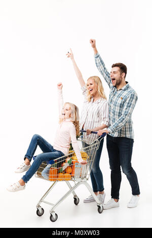 Full length portrait of a joyful family walking with a shopping trolley full of groceries isolated over white background, little girl sitting in cart Stock Photo