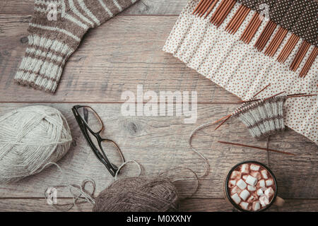 The process of knitting sock with circular bamboo knitting needles, glasses, woolen yarn of natural color, glasses on a wooden rustic table. Stock Photo