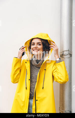 Smiling adult girl in yellow coat putting hood on head posing on camera with candid smile, being happy Stock Photo