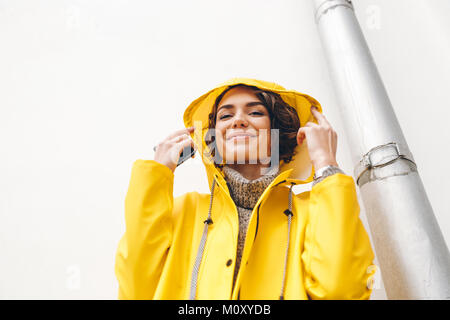 Pretty young woman with curly brown hair wearing yellow coat due to bad weather, standing in front of white wall outdoors Stock Photo