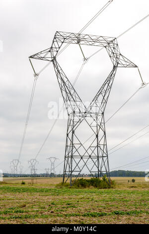 A transmission tower in the middle of a plowed field supporting high-voltage lines with dozens of other electric pylons in the background under a clou Stock Photo