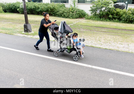 Hispanic woman pushing stroller with baby and toddler along Midtown Greenway Trail while reading her cell phone. Modern. Minneapolis Minnesota MN USA Stock Photo