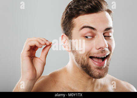 Close up portrait of a cheery man cleaning his ears with a cotton swab isolated over gray background Stock Photo