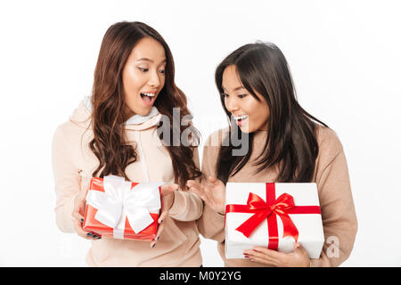 Picture of two asian cute ladies sisters standing isolated over white background holding gift boxes surprise. Looking aside. Stock Photo