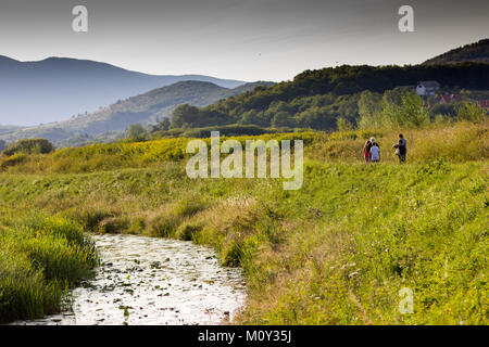 Old Gacka river passing through green mountainous countryside with small group of people (including children) walking along the grassy bank Stock Photo
