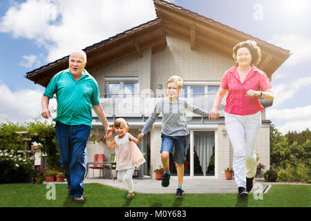 Smiling Grandparents Running With Their Grandchildren In Front Of Their House Stock Photo