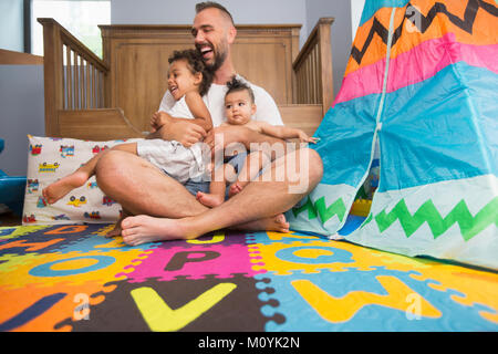 Father sitting on floor of playroom holding son and daughter Stock Photo