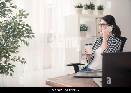 professional young female office worker using mobile cell phone calling for friend chatting when she finished work at home sitting on workplace lookin Stock Photo