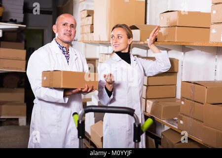 Smiling young female and male transporting cart cardboard cases in storage Stock Photo