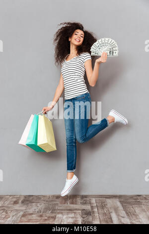 Brunette female in casual posing with a lot of packages and fan of dollar bills in hands being rich and satisfied after shopping, over grey wall in st Stock Photo