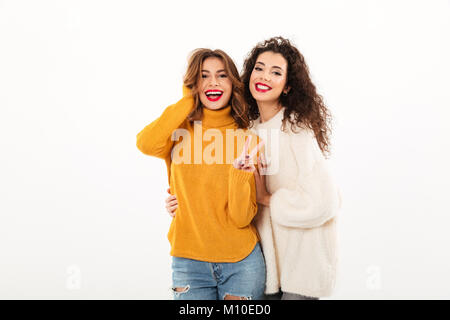 Two smiling girls in sweaters posing together and looking at the camera while one woman showing peace gesture over white background Stock Photo