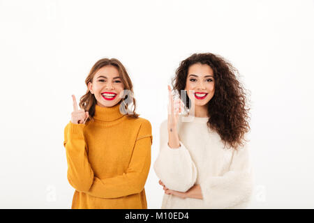 Two smiling girls in sweaters pointing up and looking at the camera over white background Stock Photo