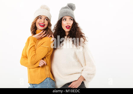 Two smiling girls in sweaters and hats standing together while looking away over white background Stock Photo