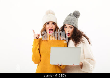 Two surprised girls in sweaters and hats standing together while using laptop computer over white background Stock Photo