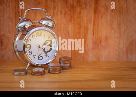 Time is Money Concept : Alarm clock put on wooden table with stack of silver money coins (Baht) in foreground. Stock Photo