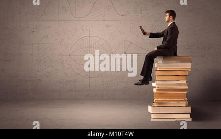 A serious businessman with tablet in hand in suit sitting on a pile of giant books in front of a greyish brown wall including drawn lines, angles, num Stock Photo