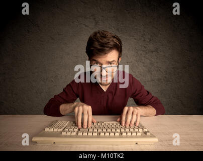 A young hacker with glasses dressed in casual clothes sitting at a desk and working on a computer keyboard in front of black clear concrete wall backg Stock Photo