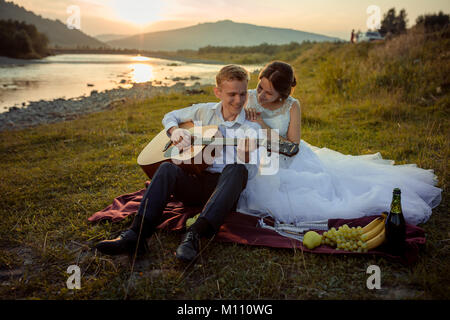 Wedding picnic composition on the river bank during the sunset. Smiling groom is playing the guitar while bride is hugging him back while sitting on the plaid. Stock Photo